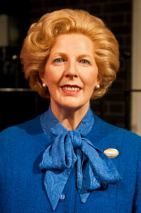 Margaret Thatcher with a styled blond hairdo, wearing a blue suit and a scarf, against a brick wall background.